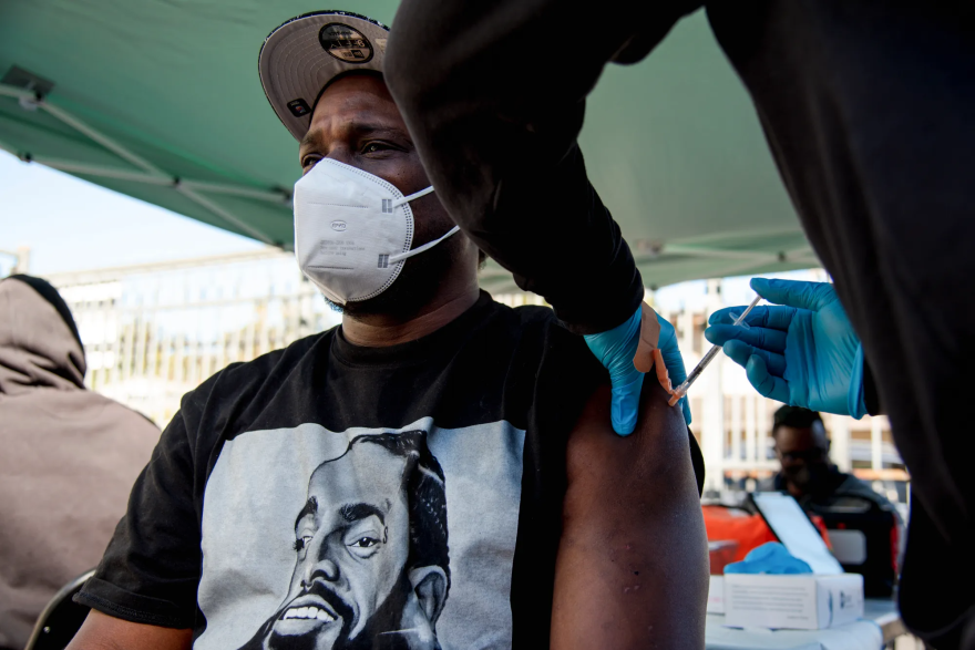 A man wearing a white mask receives a COVID vaccine injection in a facility set up under an outdoor canopy.