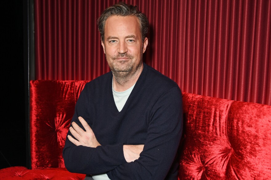 A man in a V-neck sweater with his arms crossed, sitting on a red velvet couch and smiling at the camera.