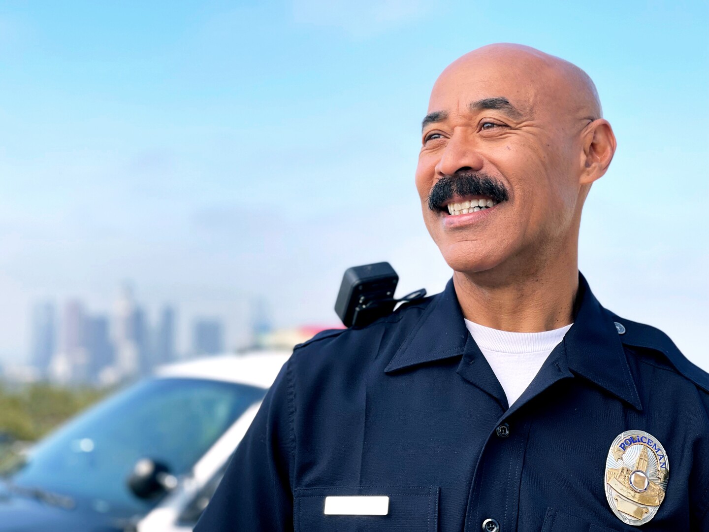 Cecil Rhambo in his LAX Police Chief uniform, looking off in the distance, with a police car behind him and the LA skyline in the far distance.
