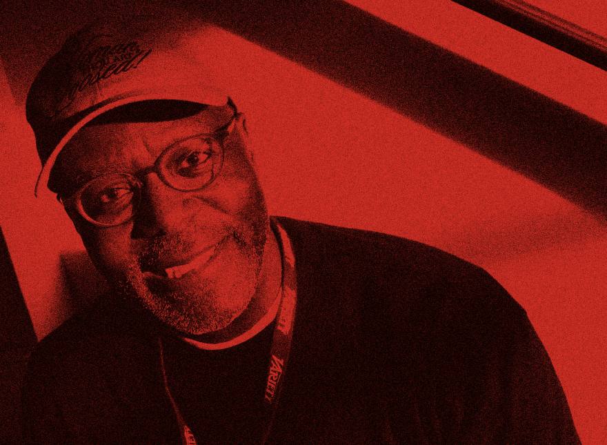 A Black man wearing glasses and a ballcap smiles at the camera as light pours in from an off-camera window.