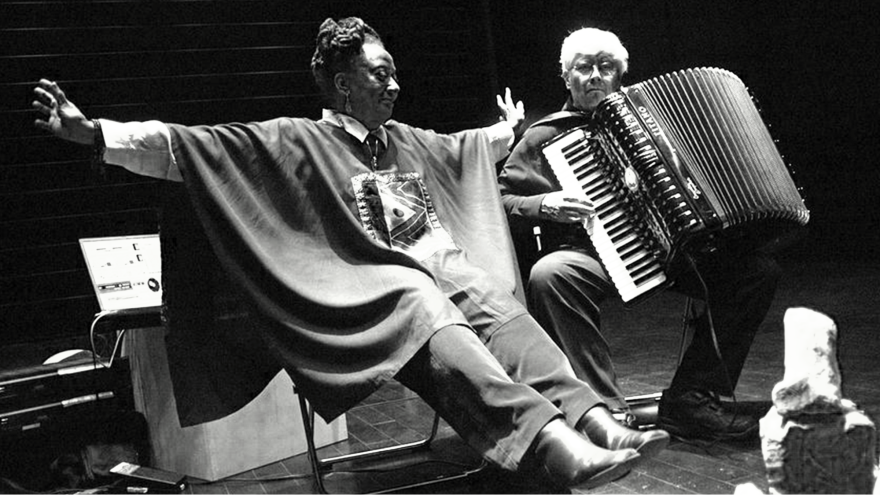 A black and white photo of two women sitting on chairs. To the right, there is a light-skinned woman holding an accordion. To the left, a dark-skinned woman holds her arms out open, wearing a large poncho.