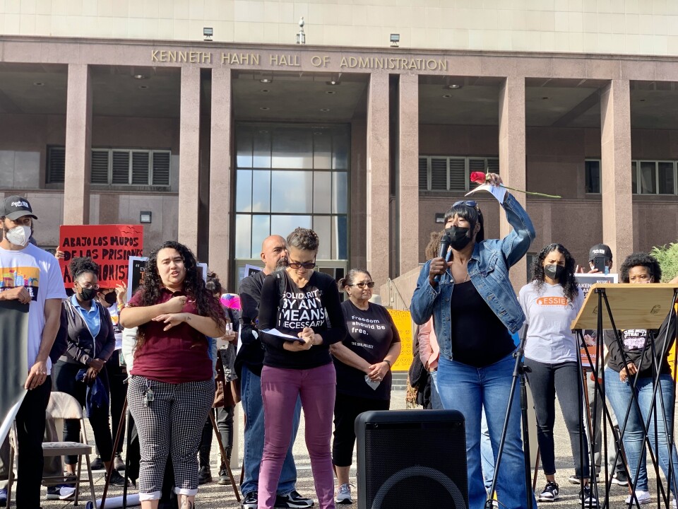Many people are gathered in front of a jail building. They are protestors. One of them, clad in a jean jacket, black shirt and jeans, speaks into a microphone.