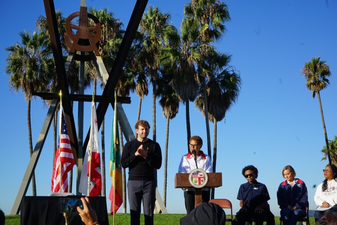 A woman stands behind a podium and a man motions beside her. Several other people stand behind them with palm trees in the distance. 