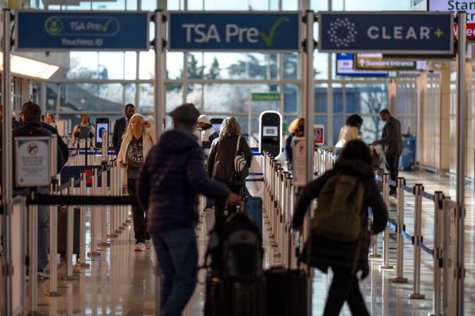 Travelers holding bags and backpacks walk through a security line in an airport with a large window in the background.