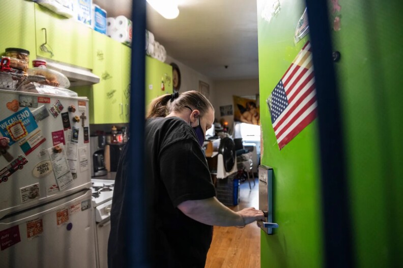 A light-skinned woman in a Black t-shirt holds the handle of an open green door. She has her brownish hair back in a ponytail. She stands in a kitchen area, a refrigerator covered with various items behind her, along with green cabinets and a stove. There are supplies stashed above the cabinets, including paper towels. There is food on top of the refrigerator. An American flag decal is on the door, which has a metal handle.