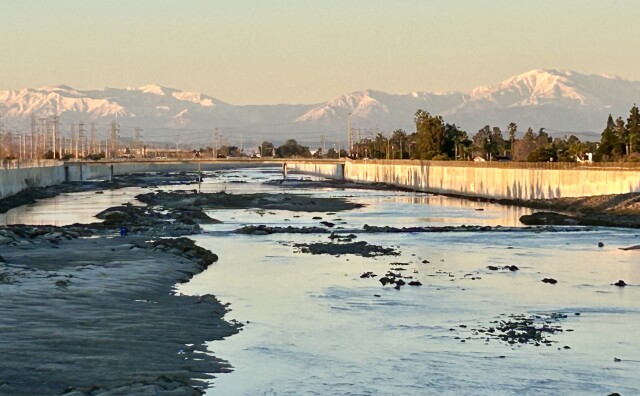 Snow capped mountains are in the distance as the golden hour sun reflects on a river with concrete embankments.