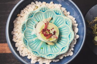 Overhead view of an oyster dish in a turquoise, scalloped ceramic dish set in a blue bowl of white stones. A pale green disc is topped with red seaweed and a small white blossom.