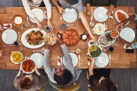 An overview of a dining table with people passing plates to one another. On the table, along with white dinner plate settings, is the typical Thanksgiving feast - a turkey, corn, pumpkin pie, ham, carrots, mashed potatoes and a large pumpkin in the middle.