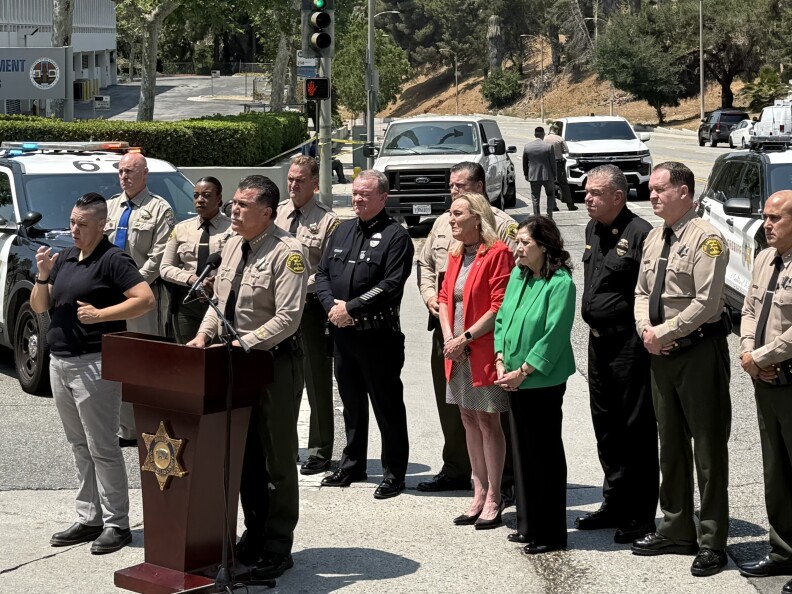 A man in a law enforcement uniform speaks at a podium. People stand behind him in law enforcement uniforms and civilian clothes.