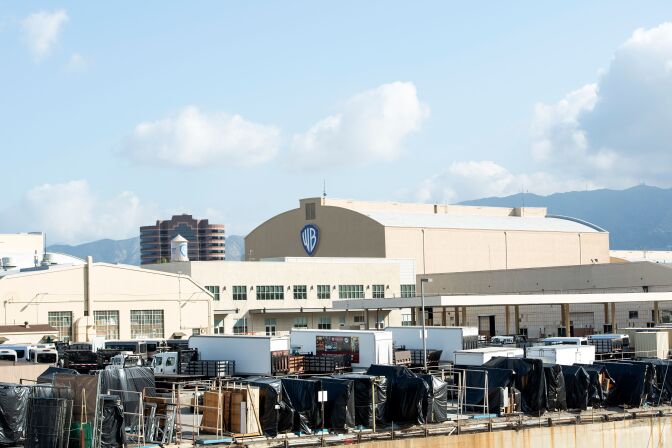 A wide view of several large beige warehouse-style buildings, one with the Warner Bros. logo. In front of the buildings are large trucks and movie equipment, mostly draped with black cloths. 
