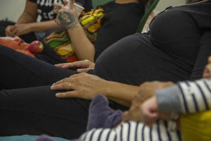 A close-up image of a pregnant woman sitting wearing a black shirt with her hands resting on her legs.