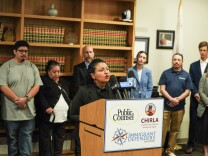 A woman speaks into microphones at a podium, with several people standing behind her.