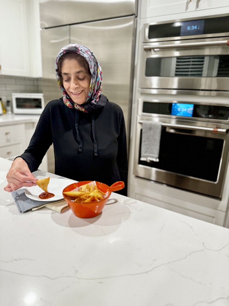 A medium skinned woman wearing a multi colored headscarf and a black top is sitting in a modern kitchen with a sleek white counter and shiny steel appliances. She's holding a samosa in her right hand and dipping it into some ketchup on a white plate. To the side is a red bowl full of samosas