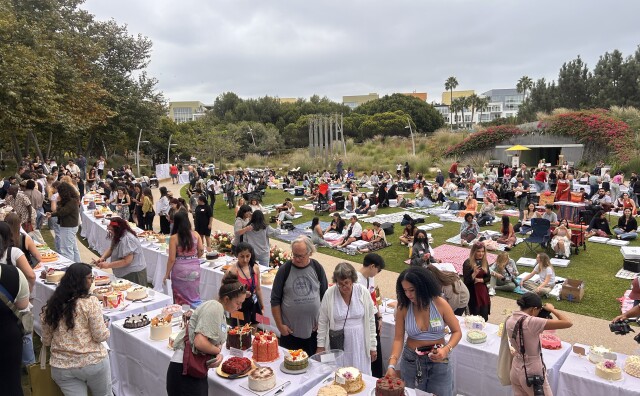 The photo depicts a lively outdoor community event centered around cakes and desserts.