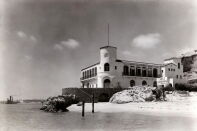 A black and white photo depicts a beachfront marine lab with a central tower and tiled roof. 