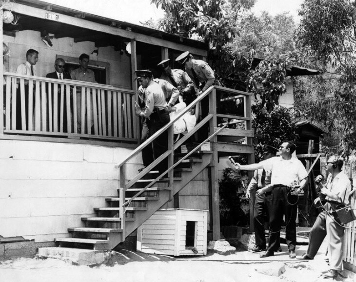 Men in uniform ddra a woman down a flight of outdoor stairs off a porch as people look on.