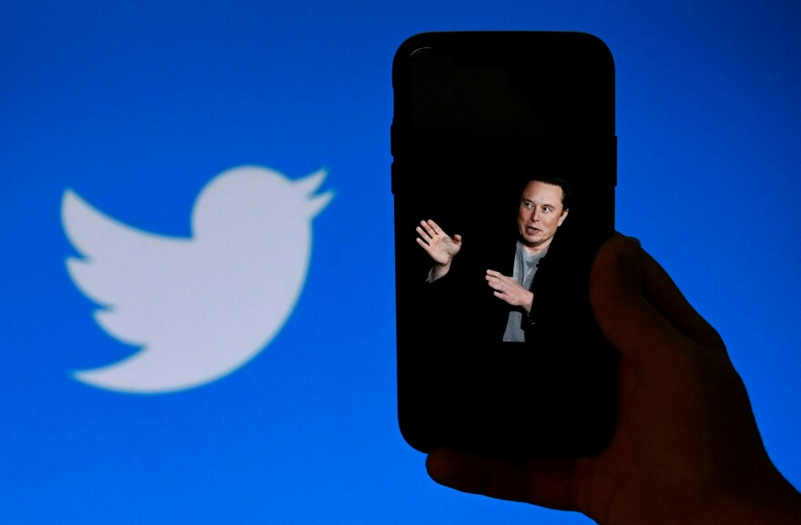 A man with light-tone skin is seening gesturing on a mobile phone toward a blue background with a white bird logo
