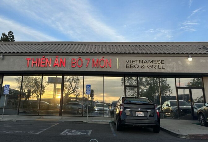 The exterior of Thien An Bò 7 Món restaurant at dusk, with a red neon sign reading "Thiên Ân Bò 7 Món / Vietnamese BBQ & Grill" above a glass storefront reflecting the sunset.