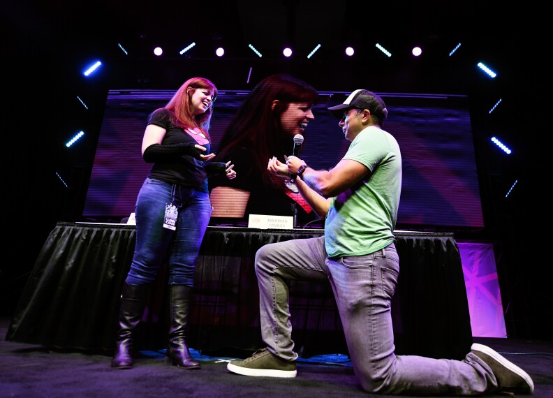 A man in jeans and a green T-shirt on one knee, holding a microphone and a ring, in front of a redheaded woman in Mary Jane cosplay and a Spider-Man T-shirt. They are on stage, with a video screen behind them focused on the woman.