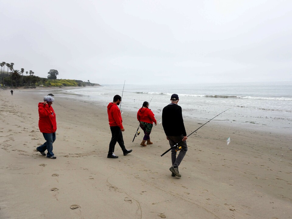 A group of four people, three of them wearing red sweatshirts, walk on the beach toward water. Their backs are to the viewer as three of the people are carrying fishing rods.