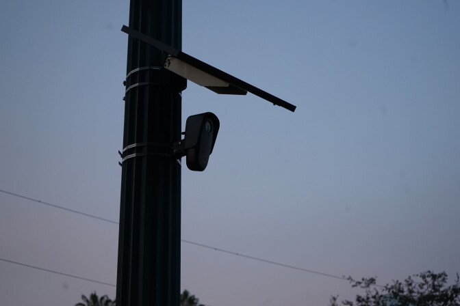 A camera is attached to a light pole, underneath a small solar panel. The sun is setting in the background and the tops of some trees are visible.