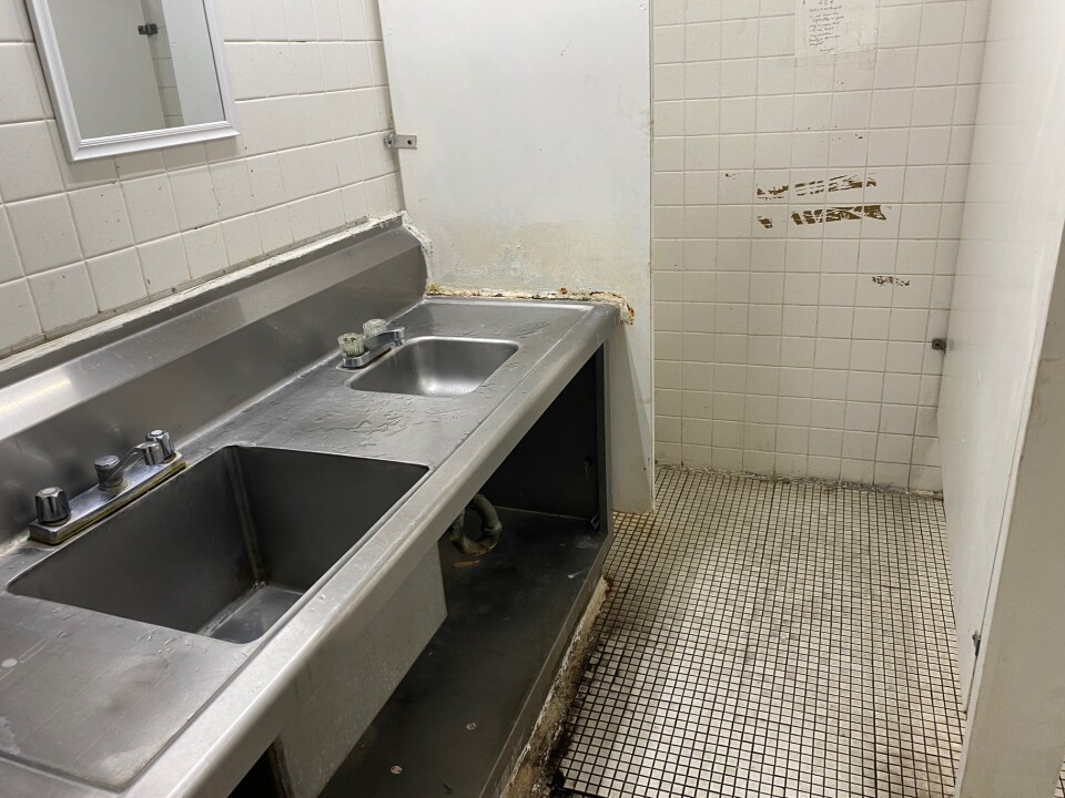 a steel double sink is affixed to the wall with a small mirror over it. The tiled floors and walls are molded and dirty.