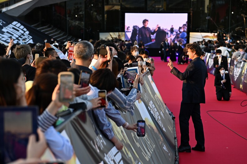 Tom Cruise waves on the red carpet to fans crowded behind barriers, many holding up phones to take pictures.