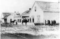 A black and white view of the U.S. Army Headquarters at Wilmington. The building is of wood and has large doors. In front of the building is a camel with two humps, a person holding the reins of a horse, a group of people standing in the front yard.