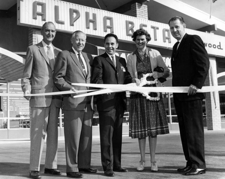A woman in a skirt holds an oversized scissors next to four men in suits in front of an Alpha Beta store.