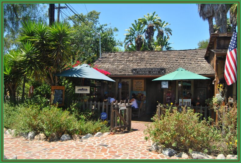 a stone sidewalk and walkway are surrounded by bushes and trees leading to a small, one-story building with tables outside and people eating at them.