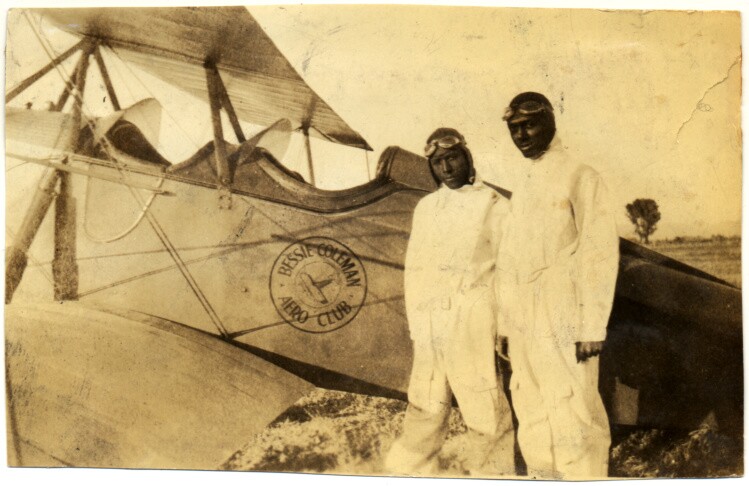 a black and white photo of two men wearing white jumpsuits, leather helmets and goggles on their head as they stand next to an old, two-seat airplane
