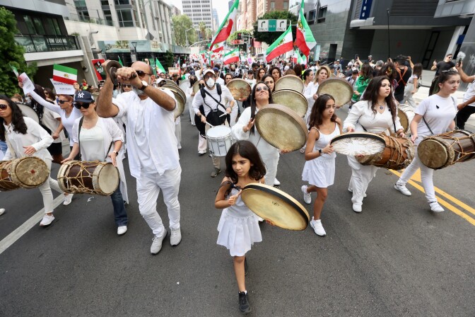 A mostly female group of drummers, women and girls, march in white on a city street.