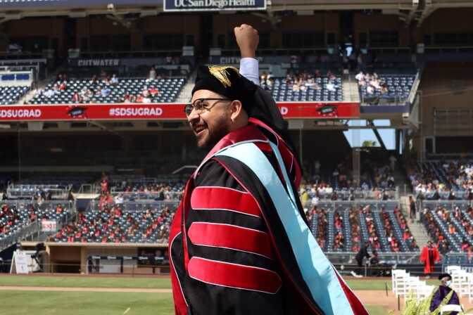 A man with glasses holds up a celebratory fist while clad in a black and red graduation gown. He also wears a black tam and light blue hood. A semi-empty stadium can be seen in the background.