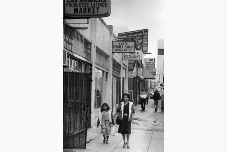 A woman and her daughter, both in dresses and coats, walk along a city street in Los Angeles. The image is in black and white.