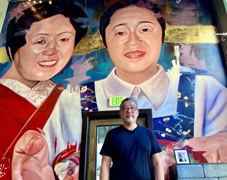 A 61-year-old Japanese American with glasses and a gray goatee stands in front of an indoors mural of two smiling Japanese American women.