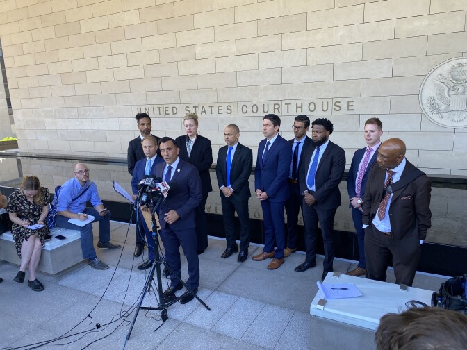 A man with light brown skin wearing a blue suit stands in front of a bank of microphones. Nine other people are standing behind him in front of a wall with the words United States Courthouse etched into it. Two reporters are seated to their left.