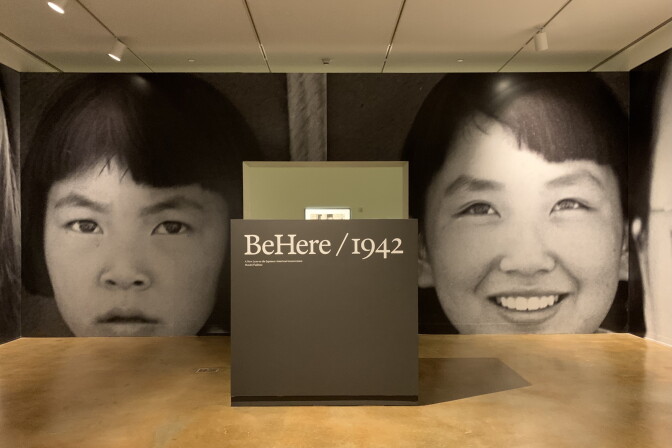 Photo of a room in a museum with giant photos of two young Japanese American girls plastered on one wall. In the foreground is a sign that reads "BeHere/1942," the name of the exhibition.