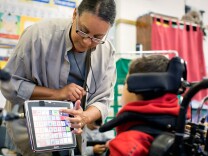 A woman in glasses helps a young boy in a wheel chair with navigating a computer screen.