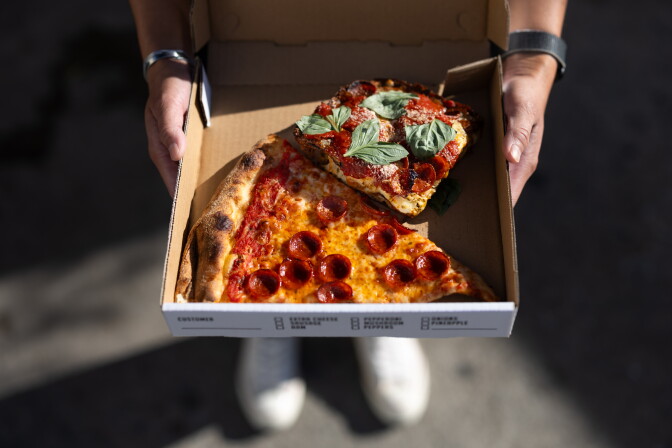 An overhead photo of two hands holding an opened cardboard pizza box: Tucked inside are a large thin crust slice of pepperoni pizza and a thick crust square slice of pizza with pepperoni and topped off with fresh basil leaves.