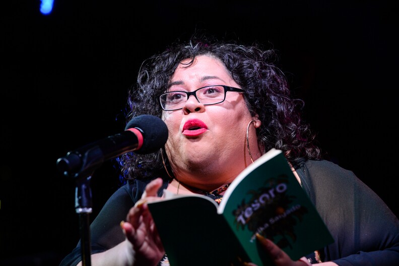 Yesika Salgado, a woman with dark curly hair and wearing glasses, speaks into a microphone while holding and reading from a book titled 'Tesoro.'