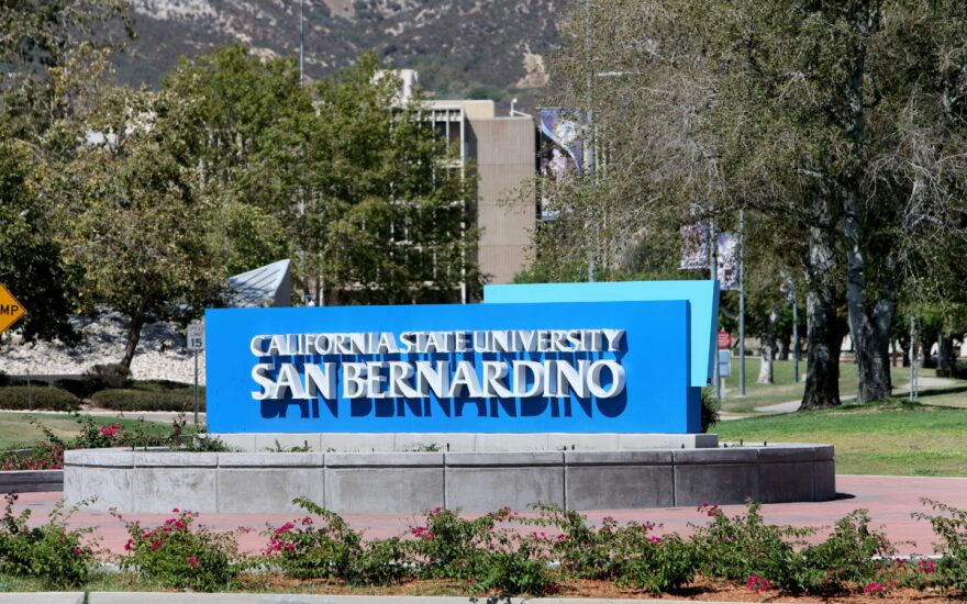 The blue California State University San Bernardino sign with a library in the background.
