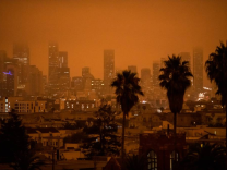 Orange-tinged skies caused by smoke enshroud a city skyline. Palm trees are in the foreground
