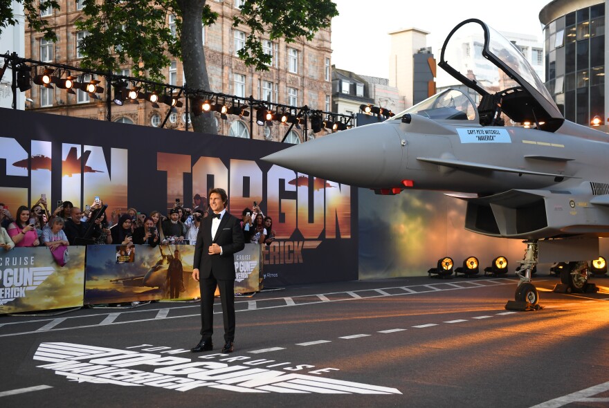 Tom Cruise in a classic tuxedo stands on a road in front of a fighter jet.