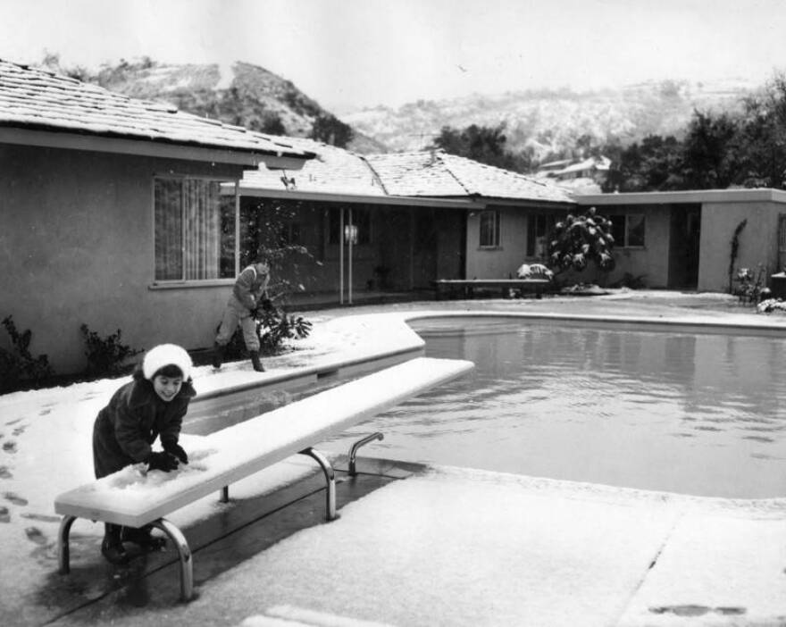 A black and white photo of two kids near a backyard pool scooping up piles of snow from the ground. One is scooping off the diving board. In the background are speckled mountains.