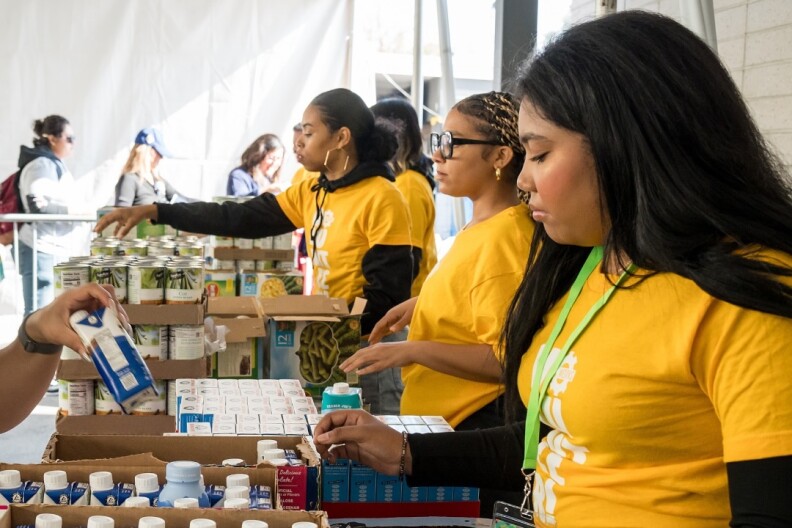 Three women wearing yellow shirts stand behind a table and hand out food and drinks to patrons. In the background, a line of people can be seen waiting.