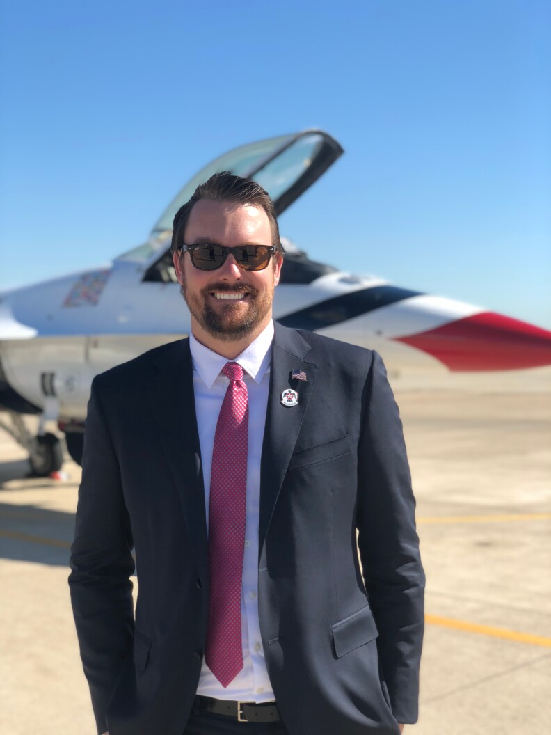 A man in sunglasses wearing a suit and red tie stands, smiling, on a tarmac with a jet plane in the background.