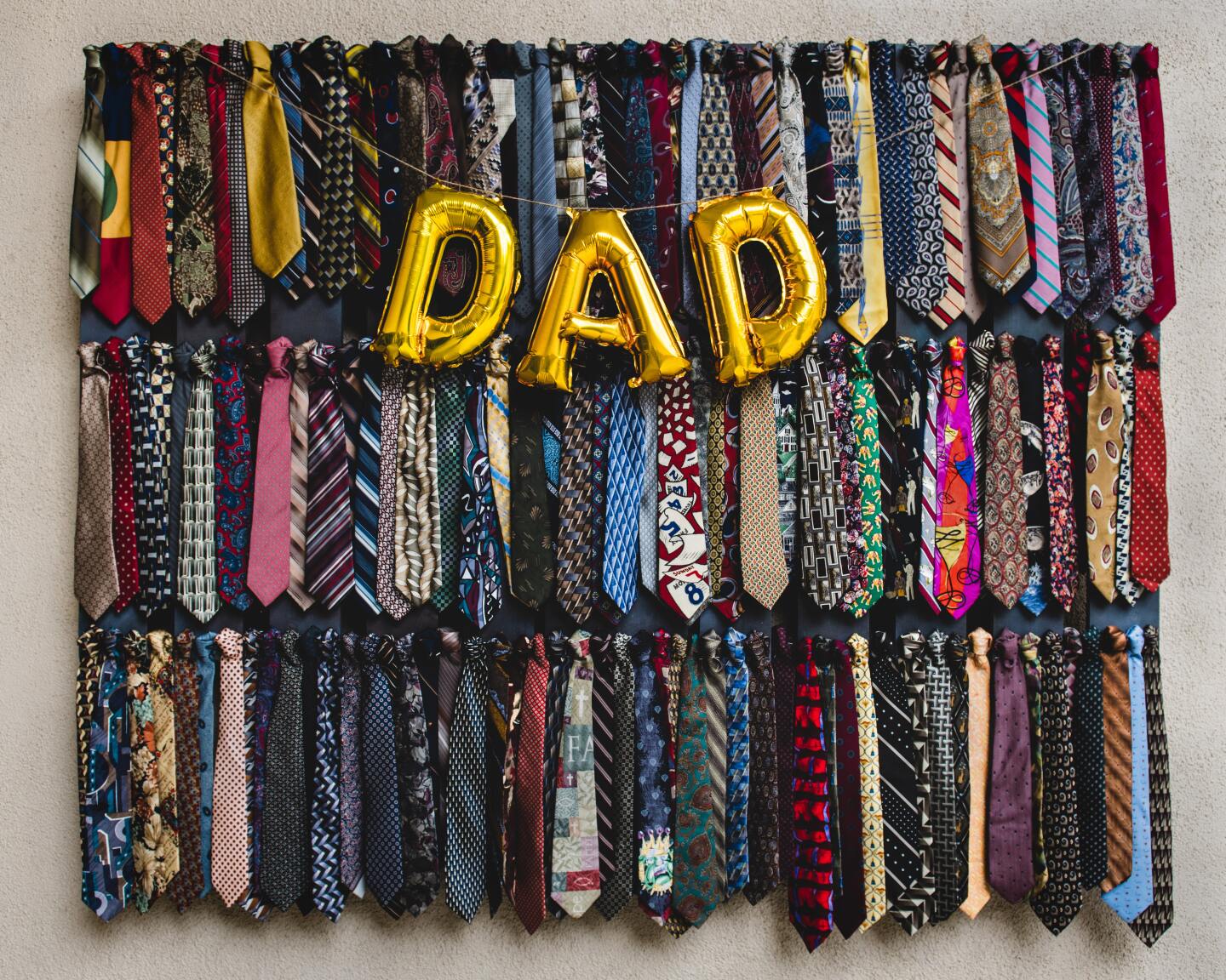 Three horizontal rows of colorful ties are overlaid with gold balloons that spell out dad in honor of father's day.