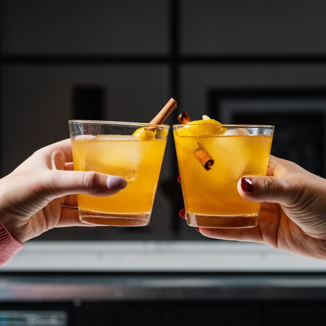 Two hands with painted nails toast yellow-orange cocktails with cinnamon sticks and lemon peel.