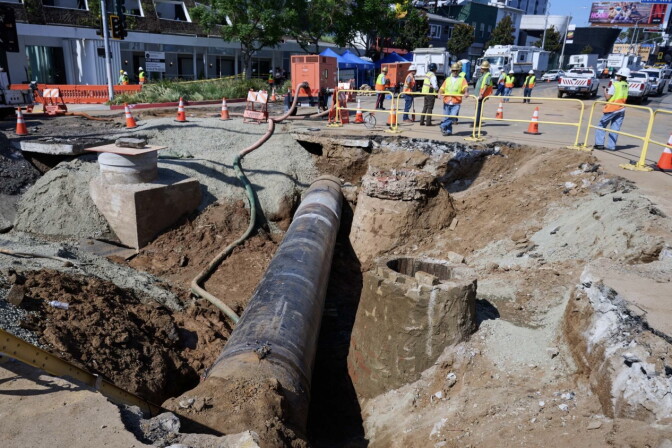 A section of a city street has been dug up, creating a deep trench with an exposed pipe running down the middle. 