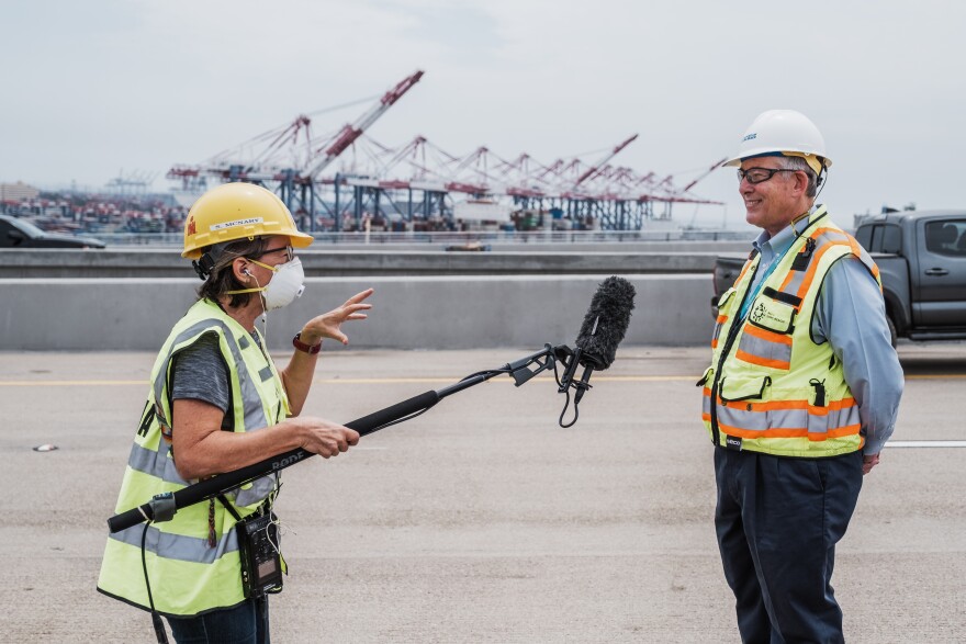 A reporter in a yellow hard hat and safety vest holds a boom mic during an interview with a man in a white hard hat and yellow safety vest. The port is behind them.